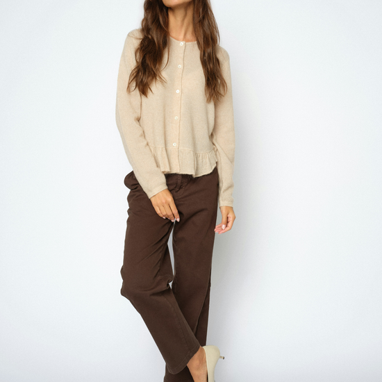 A woman modeling a light beige knit cardigan, dark brown trousers, and beige pointed-toe heels against a plain light gray studio background.