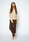 A woman modeling a light beige knit cardigan, dark brown trousers, and beige pointed-toe heels against a plain light gray studio background.