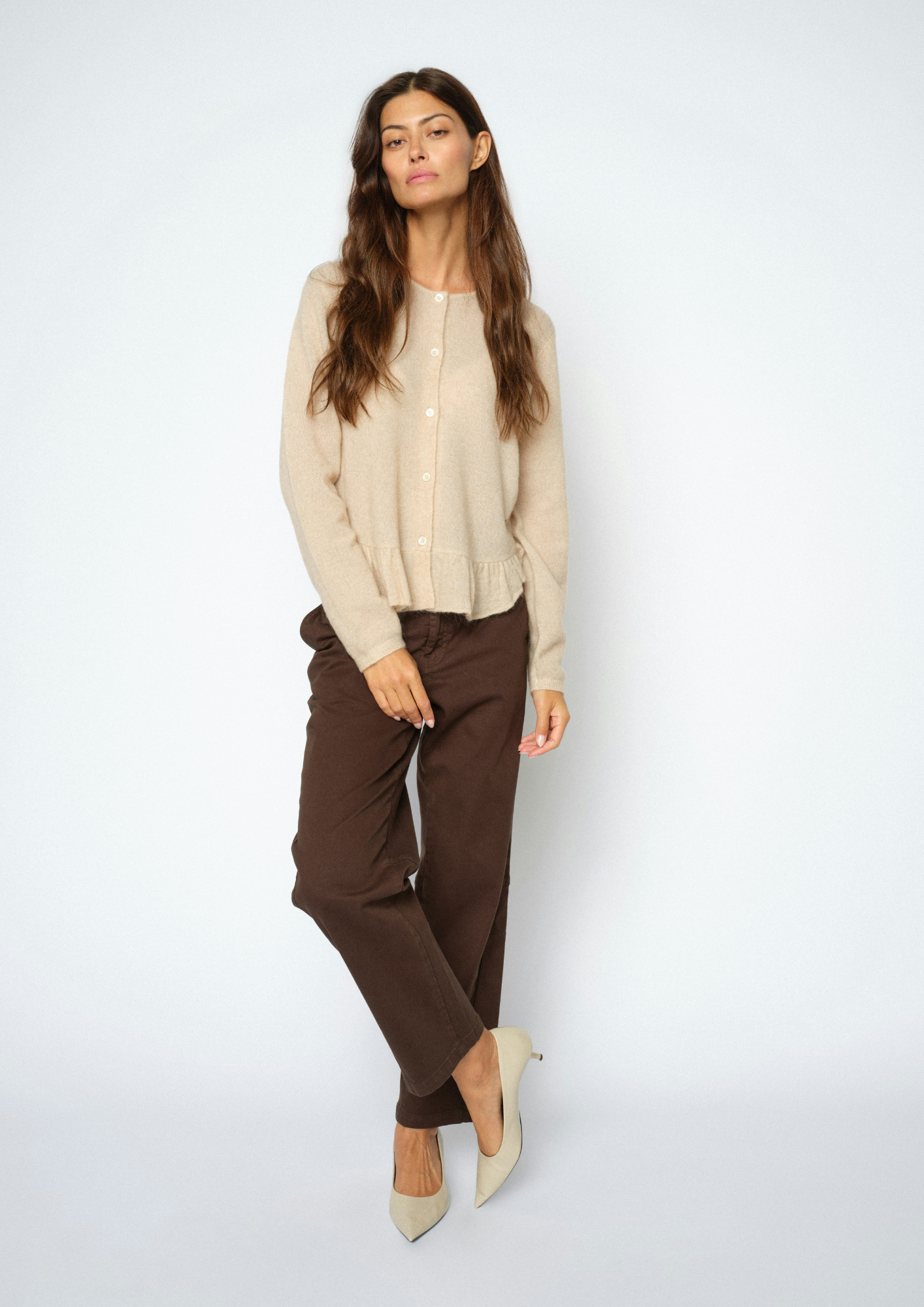 A woman modeling a light beige knit cardigan, dark brown trousers, and beige pointed-toe heels against a plain light gray studio background.