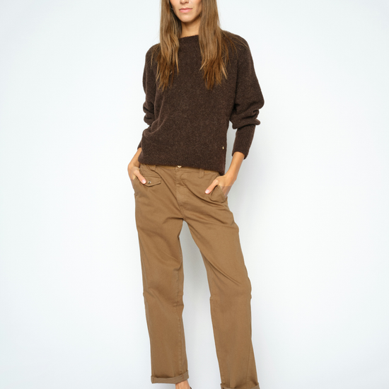 A woman modeling a dark brown knit crewneck sweater and tan straight-leg trousers with dark brown pointed-toe heels, standing against a light gray studio background.
