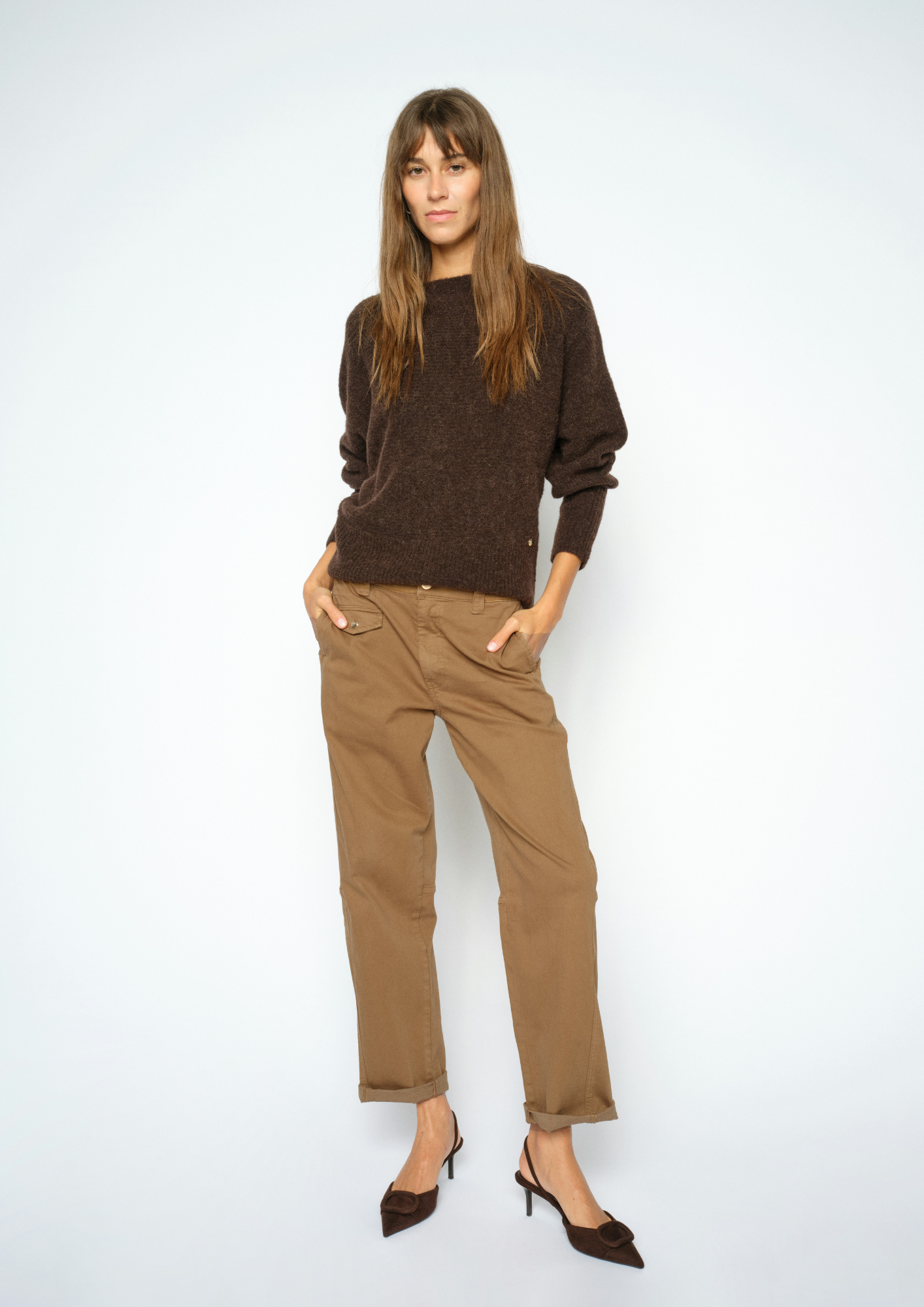 A woman modeling a dark brown knit crewneck sweater and tan straight-leg trousers with dark brown pointed-toe heels, standing against a light gray studio background.
