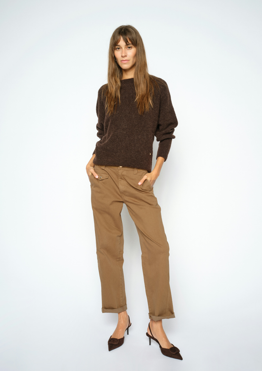 A woman modeling a dark brown knit crewneck sweater and tan straight-leg trousers with dark brown pointed-toe heels, standing against a light gray studio background.