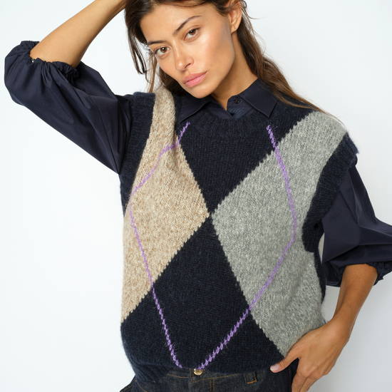 Woman wearing Blue, grey and beige Argyle vest with a blouse underneath and jeans