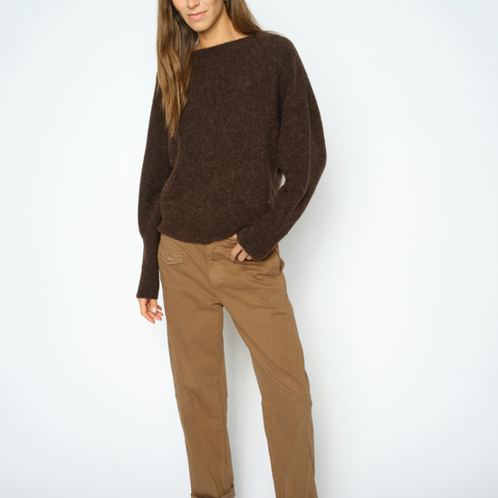 A woman modeling a dark brown knit sweater and brown trousers against a light gray studio background.