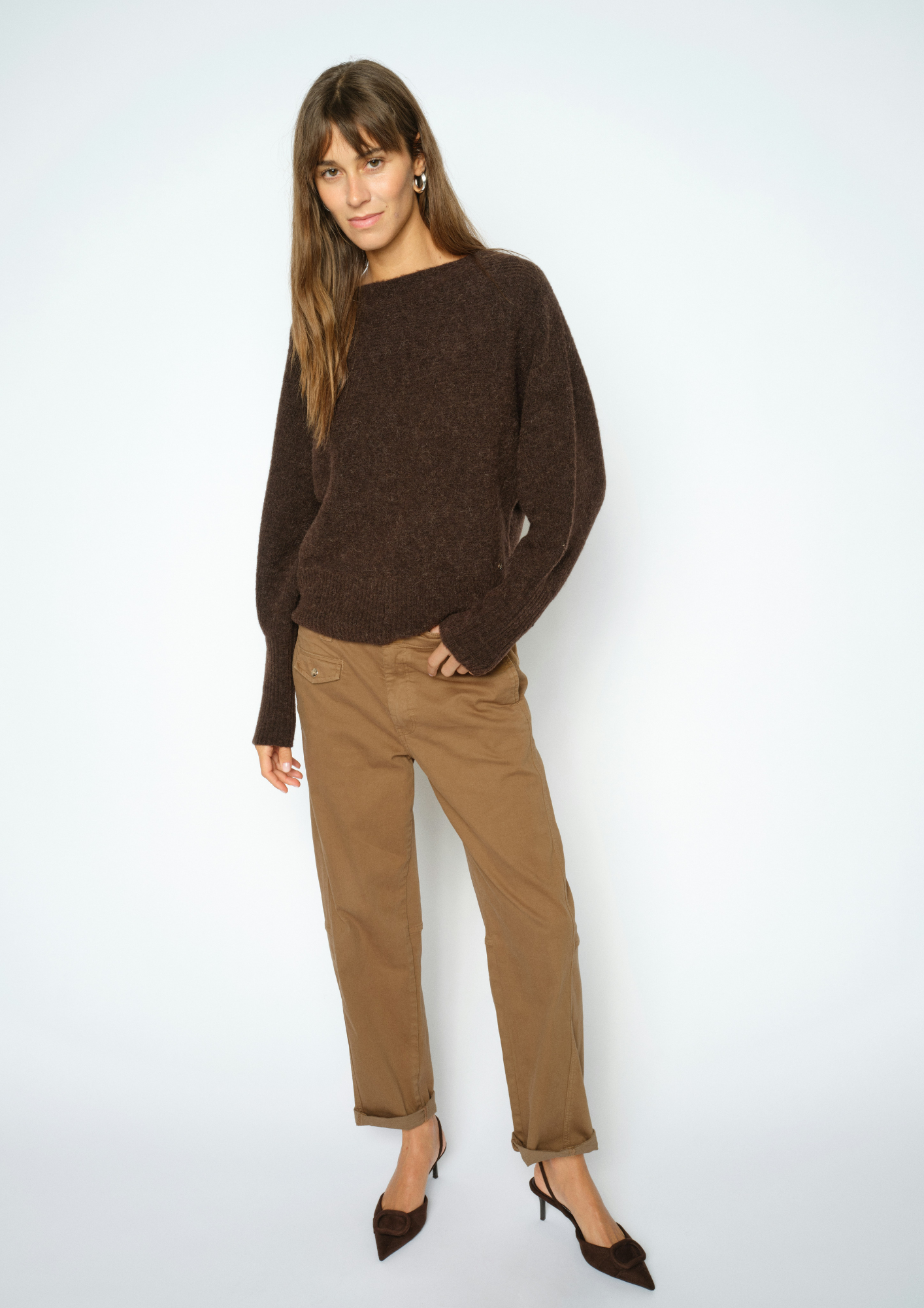 A woman modeling a dark brown knit sweater and brown trousers against a light gray studio background.