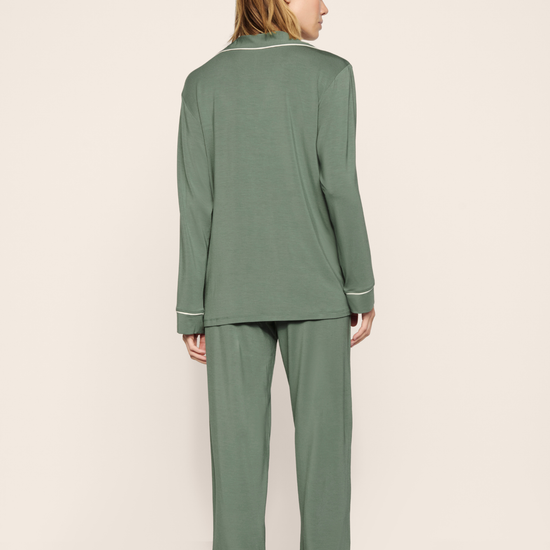 A woman shown from the back wearing a sage green two-piece pajama set with white piping details, standing against a plain beige studio background.