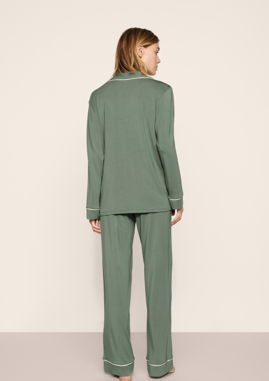 A woman shown from the back wearing a sage green two-piece pajama set with white piping details, standing against a plain beige studio background.
