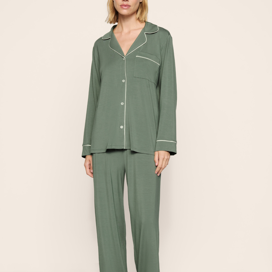 A woman modeling a two-piece sage green pajama set with white piping details, consisting of a long-sleeve button-up top and matching wide-leg pants, against a neutral beige studio background.