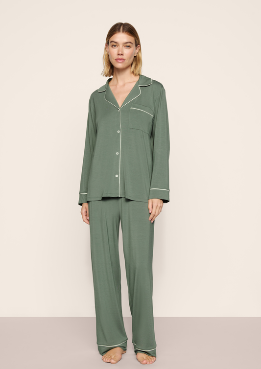 A woman modeling a two-piece sage green pajama set with white piping details, consisting of a long-sleeve button-up top and matching wide-leg pants, against a neutral beige studio background.