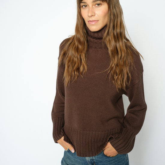A woman with long brown hair wearing a ribbed brown turtleneck sweater and blue jeans, standing against a plain white studio background.