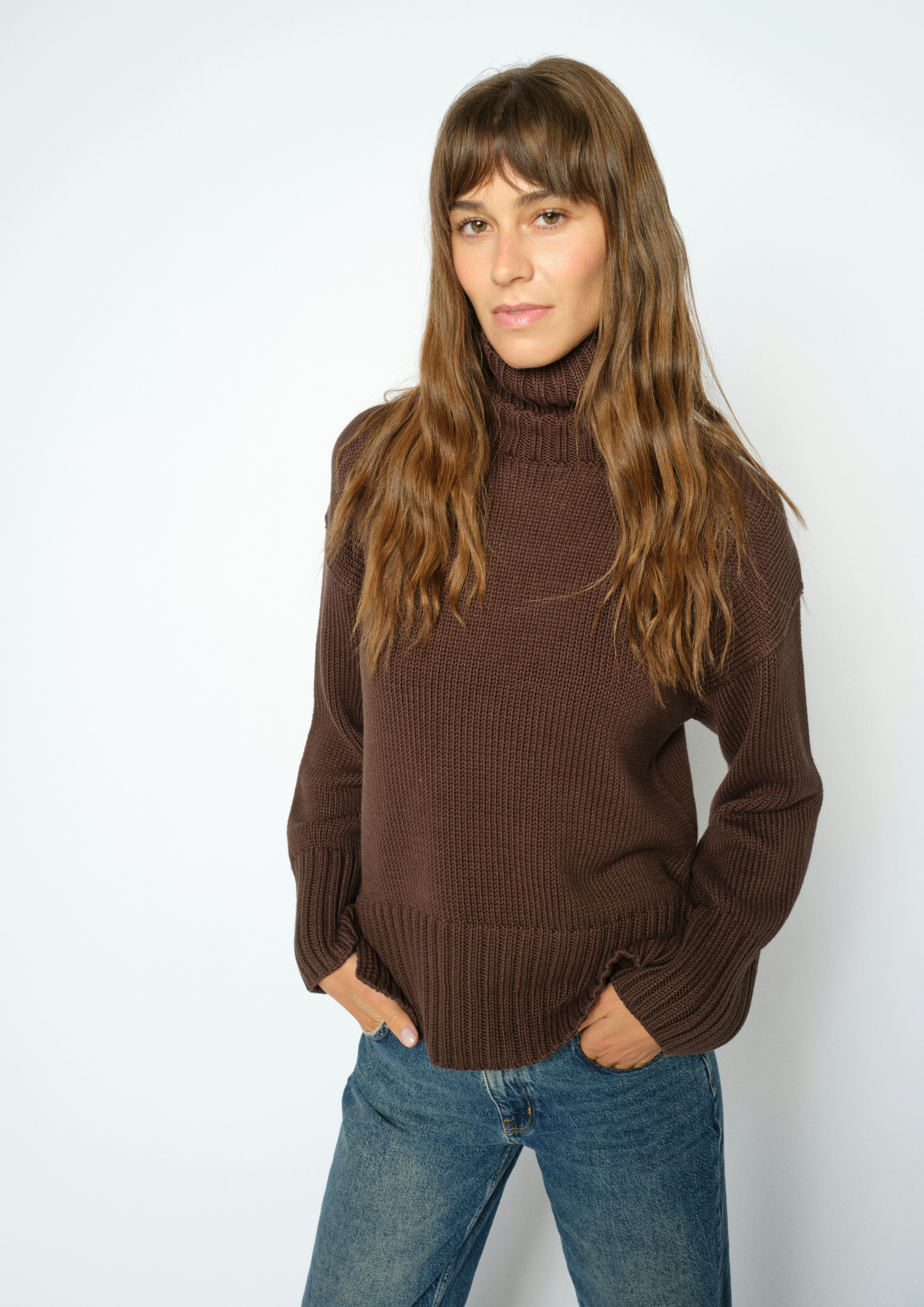 A woman with long brown hair wearing a ribbed brown turtleneck sweater and blue jeans, standing against a plain white studio background.