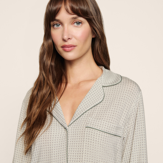 A woman with long brown hair and bangs wearing a light beige and white patterned pajama top with dark green piping, photographed against a solid light beige background.