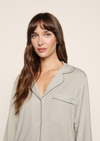 A woman with long brown hair and bangs wearing a light beige and white patterned pajama top with dark green piping, photographed against a solid light beige background.