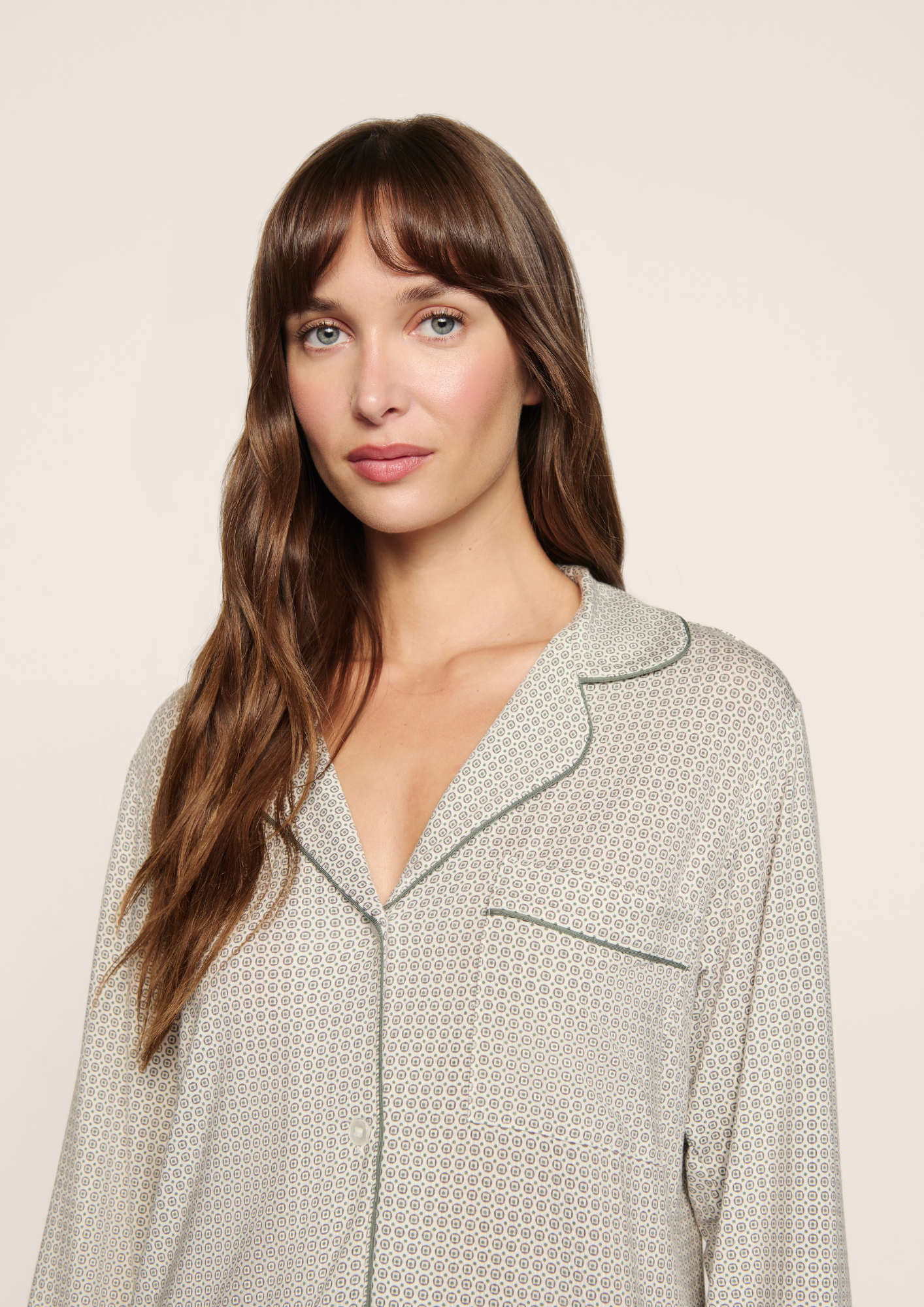 A woman with long brown hair and bangs wearing a light beige and white patterned pajama top with dark green piping, photographed against a solid light beige background.