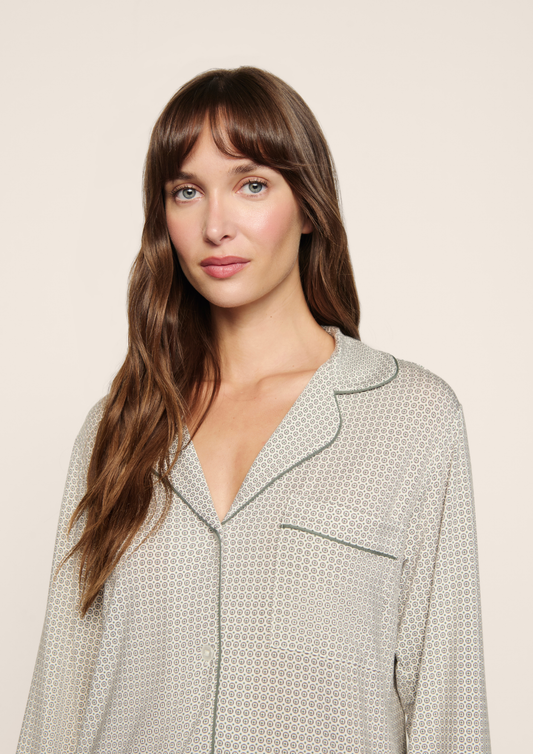 A woman with long brown hair and bangs wearing a light beige and white patterned pajama top with dark green piping, photographed against a solid light beige background.
