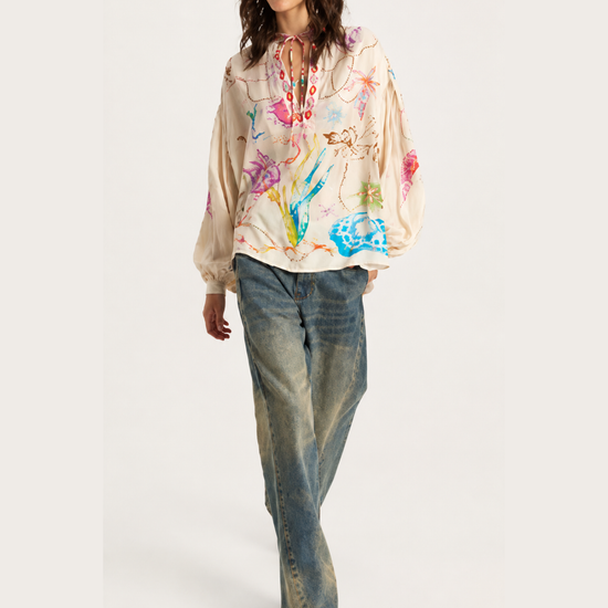 A model wearing a cream-colored hooded sweatshirt with a vibrant multicolor abstract paint splatter print, paired with blue jeans and dark brown boots, set against a plain light gray studio background.