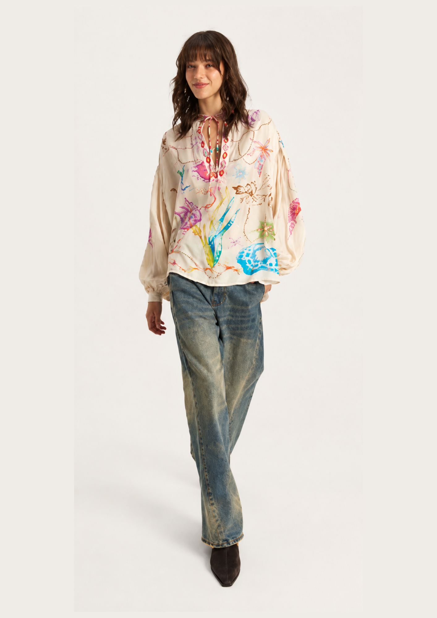 A model wearing a cream-colored hooded sweatshirt with a vibrant multicolor abstract paint splatter print, paired with blue jeans and dark brown boots, set against a plain light gray studio background.