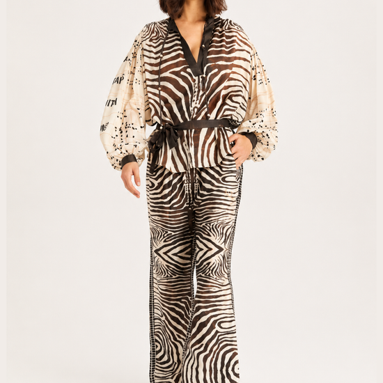 A woman modeling a matching two-piece jumpsuit with a zebra print pattern, featuring a beige and black design with a dark brown belt, photographed against a plain light gray background.