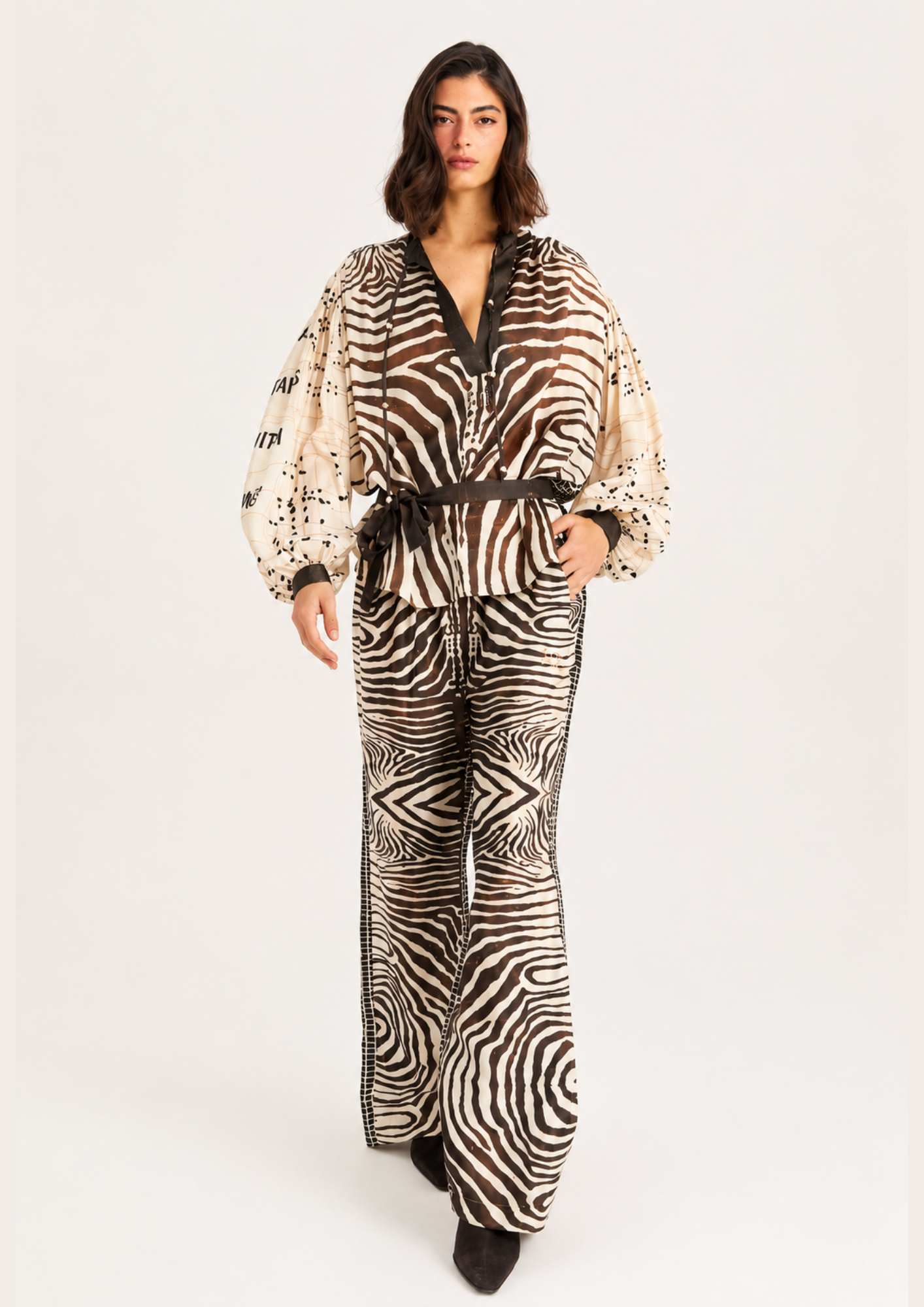 A woman modeling a matching two-piece jumpsuit with a zebra print pattern, featuring a beige and black design with a dark brown belt, photographed against a plain light gray background.