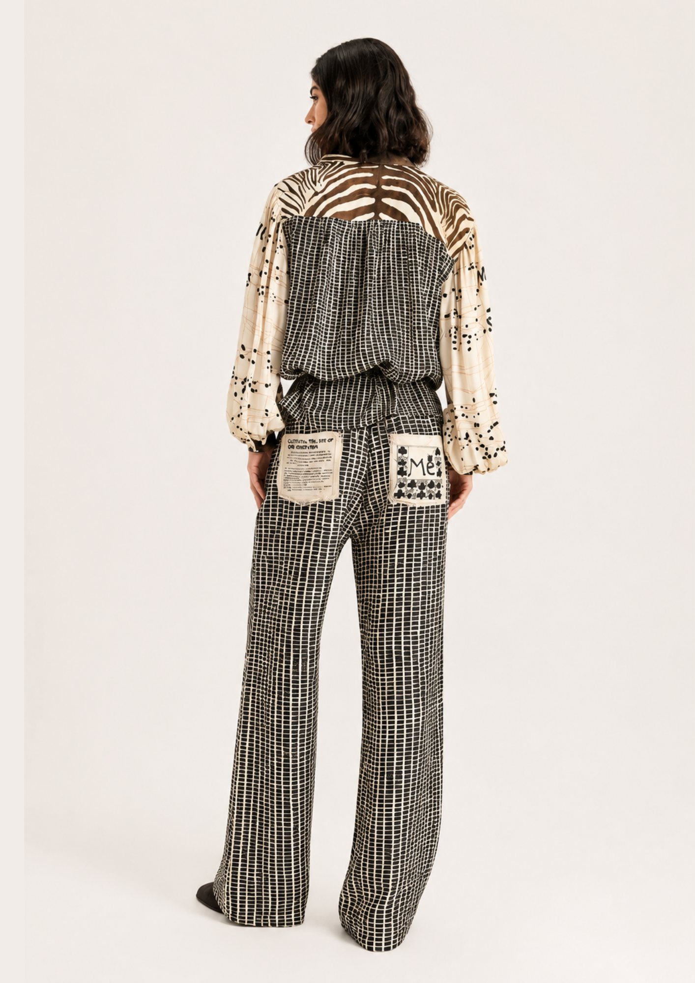 A model viewed from the back wearing a patterned jumpsuit with a cream and black-and-white houndstooth pattern, wide-leg trousers, and a back design featuring a zebra print and patch pockets.