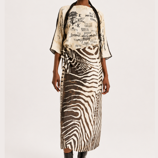 A model stands against a light gray studio background wearing a matching two-piece outfit with a cream top featuring black graphic text and a zebra-print skirt in black and beige.
