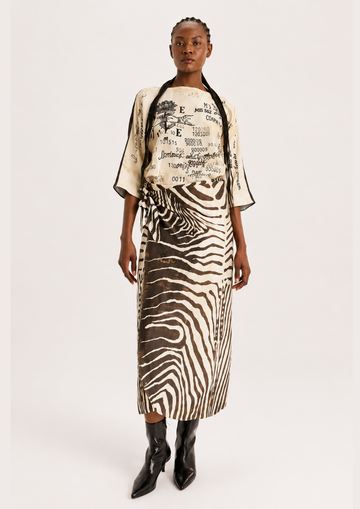 A model stands against a light gray studio background wearing a matching two-piece outfit with a cream top featuring black graphic text and a zebra-print skirt in black and beige.