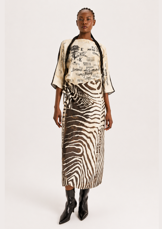 A model stands against a light gray studio background wearing a matching two-piece outfit with a cream top featuring black graphic text and a zebra-print skirt in black and beige.