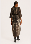 A woman viewed from the back wearing a sheer brown kimono-style dress with a zebra-stripe pattern and black ankle boots, photographed against a plain light background.