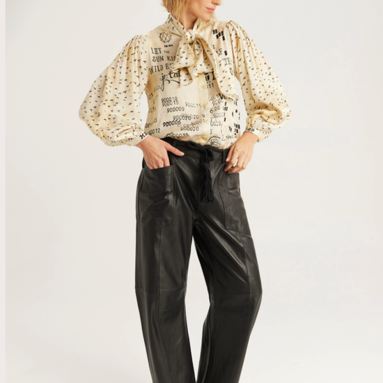 A model wearing a cream-colored blouse with black text and dot patterns, paired with black wide-leg faux leather pants and black pointed-toe shoes, against a plain light gray studio background.