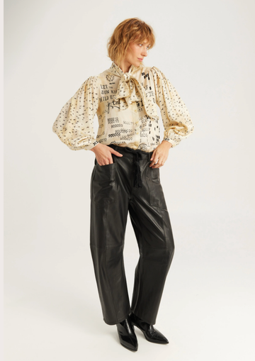 A model wearing a cream-colored blouse with black text and dot patterns, paired with black wide-leg faux leather pants and black pointed-toe shoes, against a plain light gray studio background.