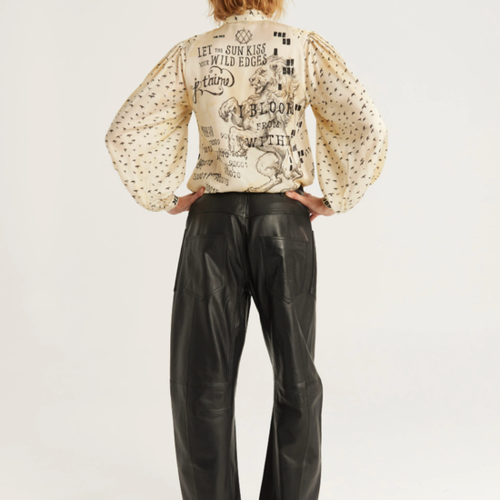 A model viewed from the back wearing a cream-colored long-sleeve blouse with black text and graphic prints, paired with black wide-leg leather pants and black heels, against a plain white studio background.