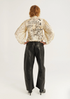 A model viewed from the back wearing a cream-colored long-sleeve blouse with black text and graphic prints, paired with black wide-leg leather pants and black heels, against a plain white studio background.