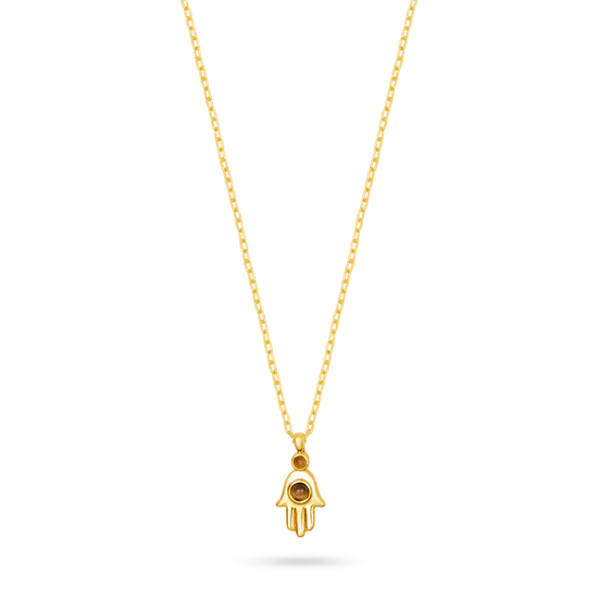 A gold Hamsa hand pendant necklace with a small black eye detail, suspended from a delicate gold chain against a solid white background.