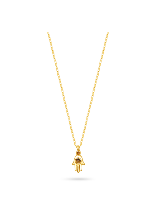 A gold Hamsa hand pendant necklace with a small black eye detail, suspended from a delicate gold chain against a solid white background.