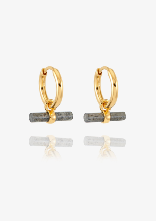 A pair of gold huggie hoop earrings with a small horizontal gray stone bar dangling from the bottom, shown on a clean white background with a soft reflection below.