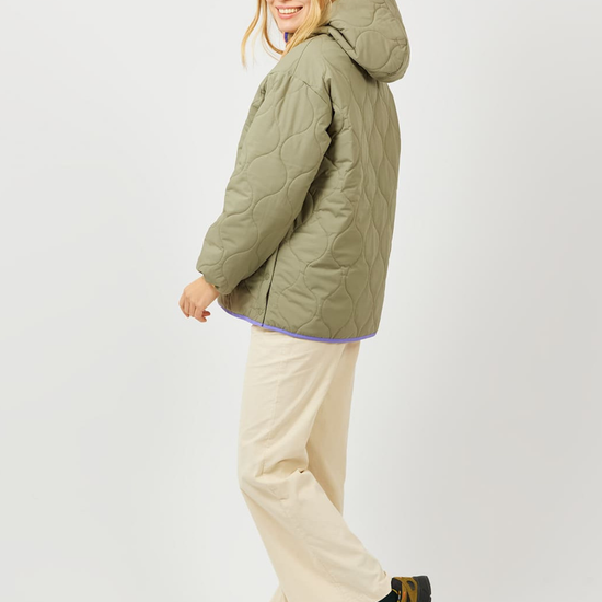 A woman modeling a quilted olive green hooded puffer jacket, paired with cream pants and patterned hiking boots, against a plain light gray studio background.