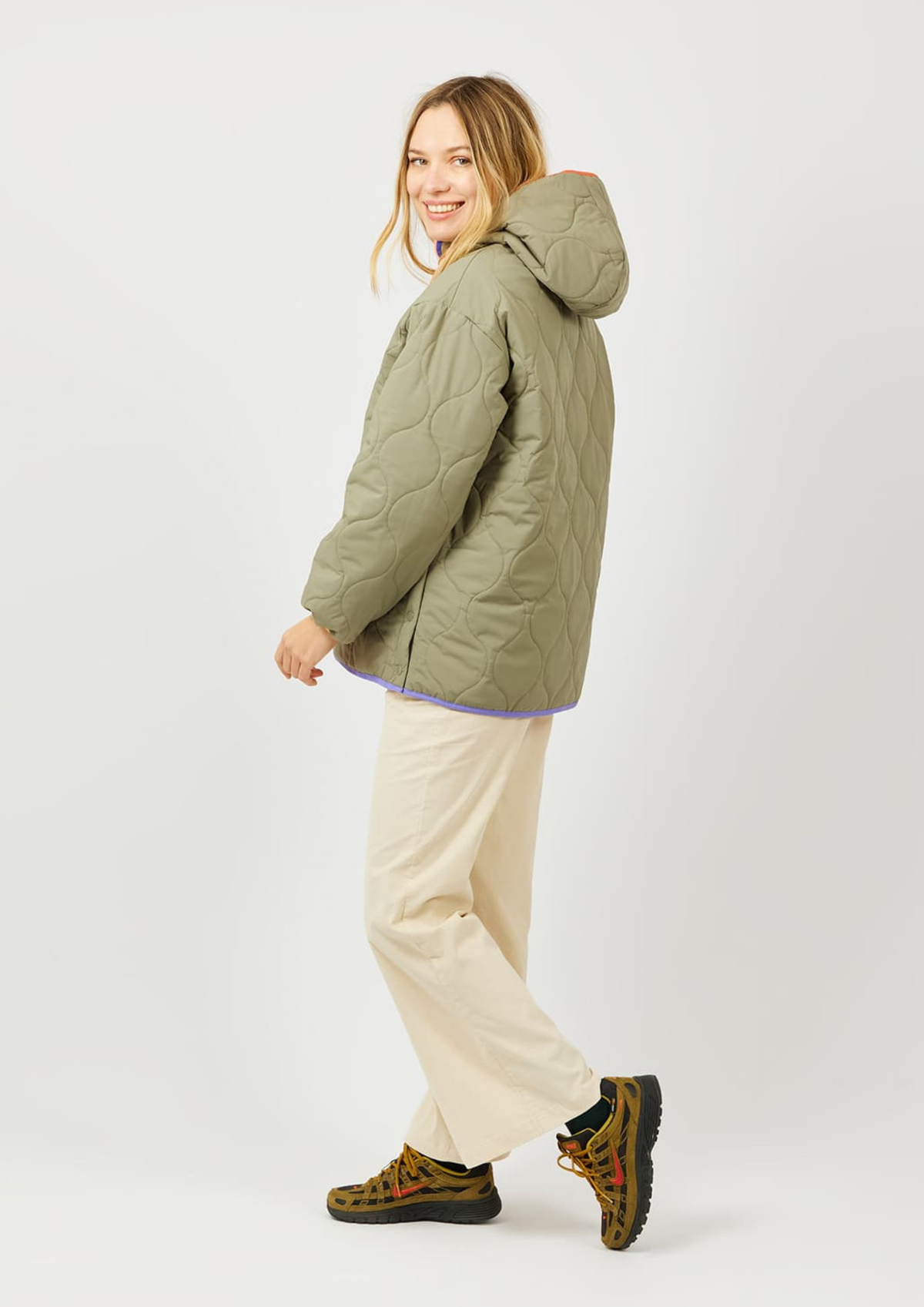 A woman modeling a quilted olive green hooded puffer jacket, paired with cream pants and patterned hiking boots, against a plain light gray studio background.