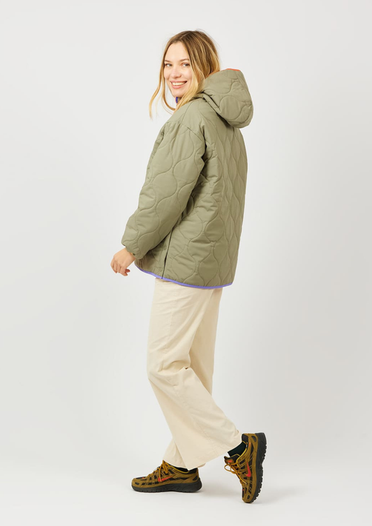 A woman modeling a quilted olive green hooded puffer jacket, paired with cream pants and patterned hiking boots, against a plain light gray studio background.