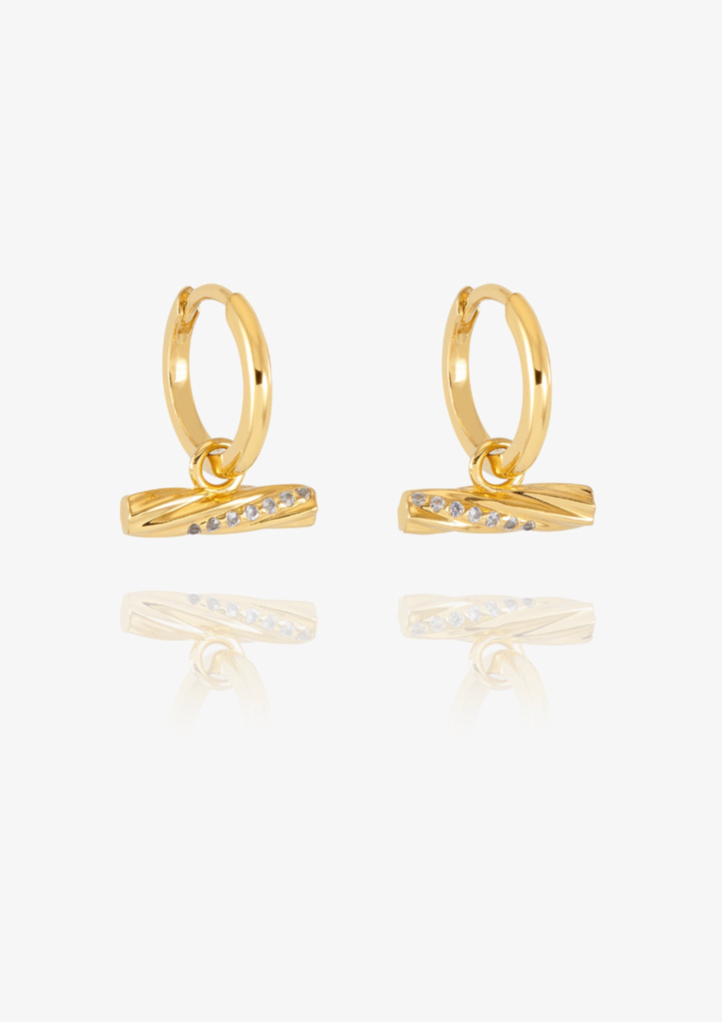 A pair of gold huggie hoop earrings with a twisted bar element set with small clear stones, displayed on a clean white background with a soft reflection below.
