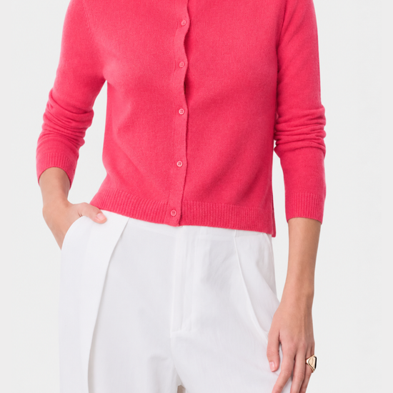 A woman wearing a bright pink knit cardigan sweater with white trousers, shown from the chin down against a clean white background.