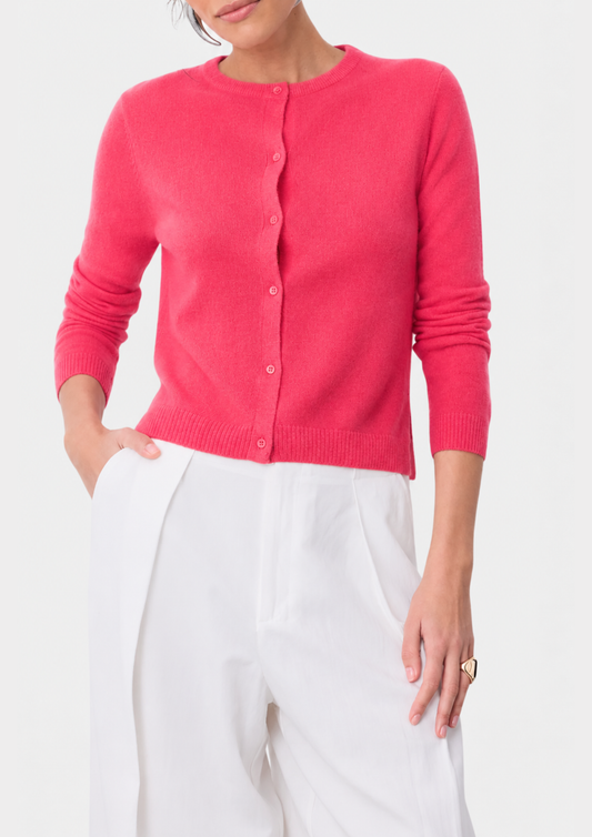A woman wearing a bright pink knit cardigan sweater with white trousers, shown from the chin down against a clean white background.