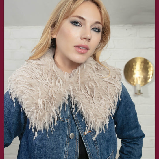 Woman wearing a denim jacket with a feathered collar against a white brick wall.