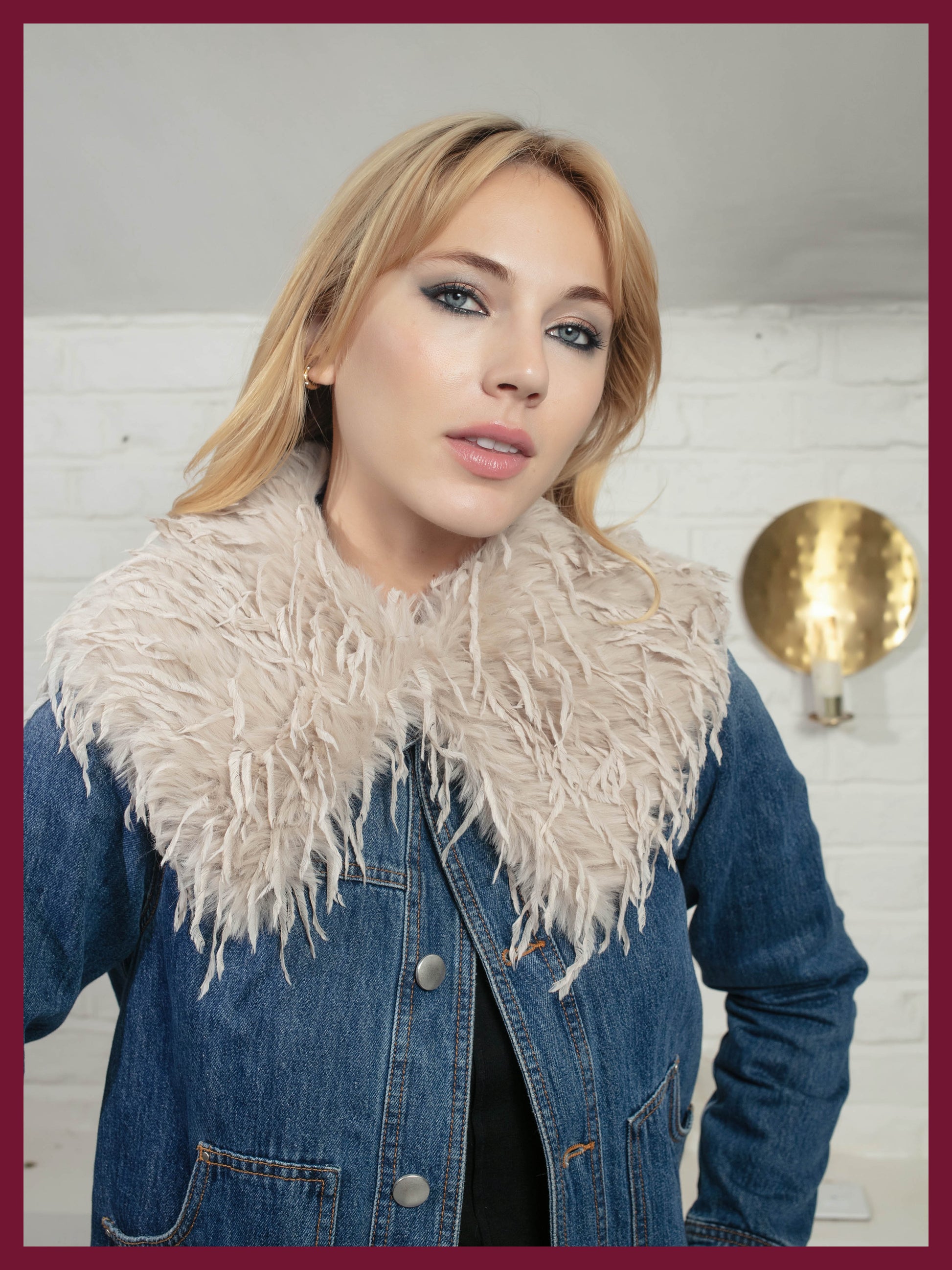 Woman wearing a denim jacket with a feathered collar against a white brick wall.