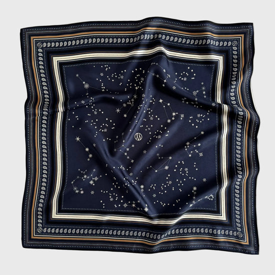 A square navy blue silk scarf featuring a celestial print of stars and constellations, framed by a decorative border with cream, gold, and navy stripes, laid flat on a light gray background.