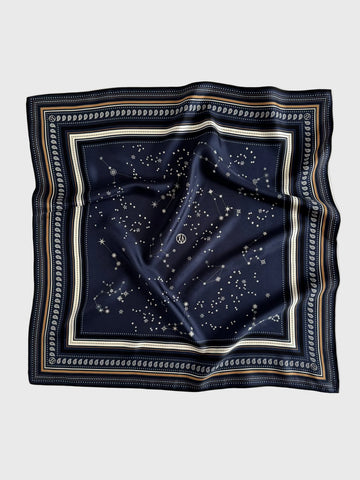 A square navy blue silk scarf featuring a celestial print of stars and constellations, framed by a decorative border with cream, gold, and navy stripes, laid flat on a light gray background.