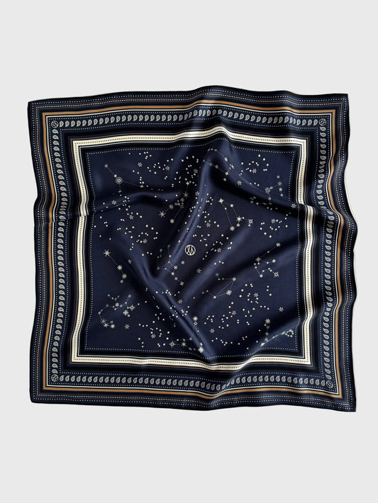 A square navy blue silk scarf featuring a celestial print of stars and constellations, framed by a decorative border with cream, gold, and navy stripes, laid flat on a light gray background.