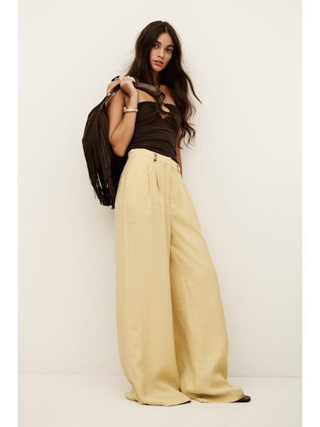 Woman wearing a black top and pale yellow wide-leg pants holding a black jacket against a white background