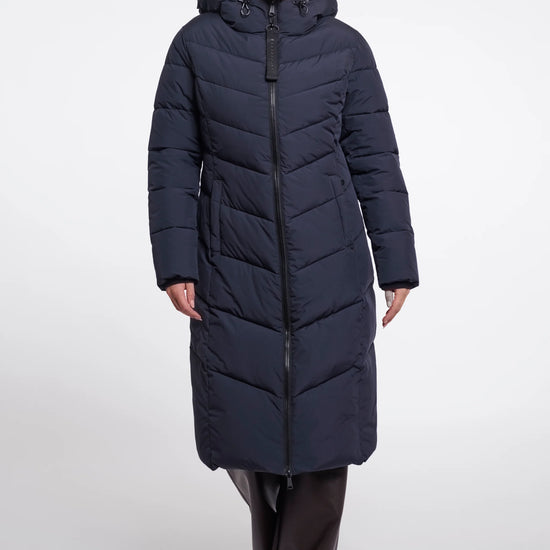 A woman modeling a long navy blue puffer coat with a hood and front zipper, paired with dark brown trousers and black shoes, against a light gray studio background.