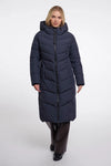 A woman modeling a long navy blue puffer coat with a hood and front zipper, paired with dark brown trousers and black shoes, against a light gray studio background.