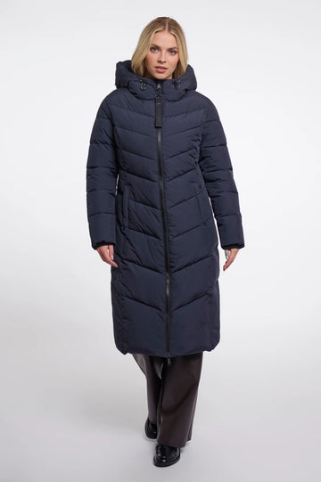 A woman modeling a long navy blue puffer coat with a hood and front zipper, paired with dark brown trousers and black shoes, against a light gray studio background.